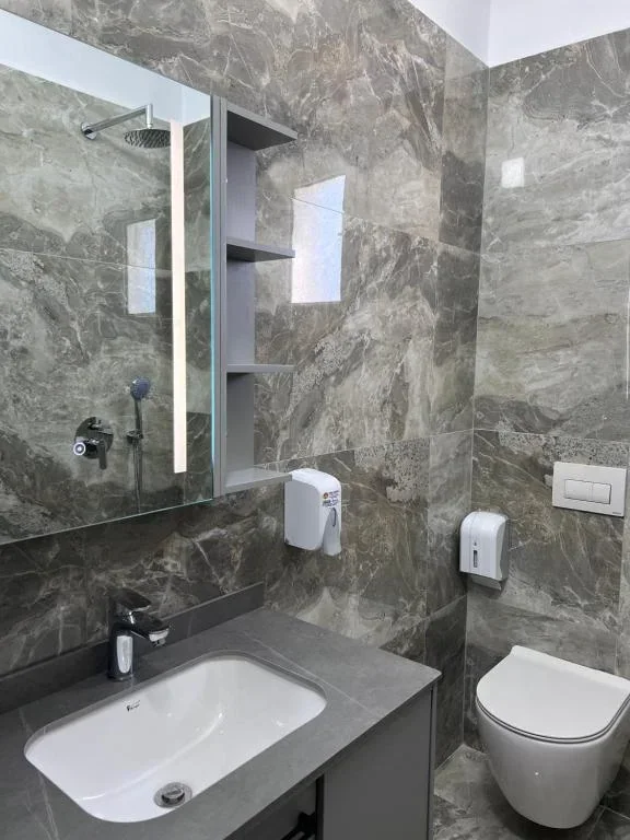 The grey marble bathroom of the Standard Quadruple Room: a lit mirror with the shower reflected in it, an open shelf unit and a wall-hung toilet