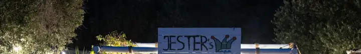 The hand-painted JESTER's sign, a blue plank board with the jester's-cap mark, standing above a rope-lashed white timber beam and lit at night