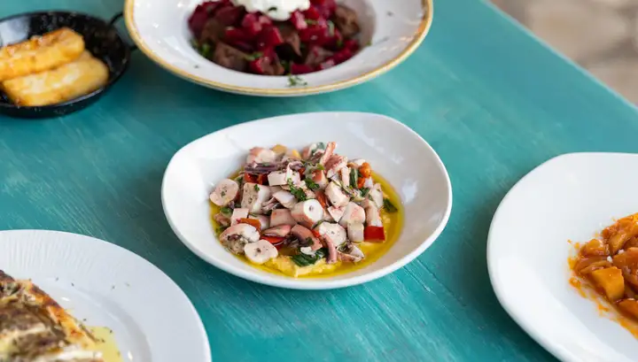A white bowl of chopped octopus with herbs, red pepper and olive oil over a purée, on a turquoise painted table