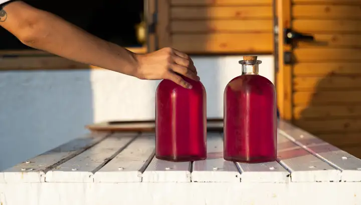 Two glass bottles of a deep crimson drink, one of them stoppered with a cork, set on a white timber counter in late afternoon sun