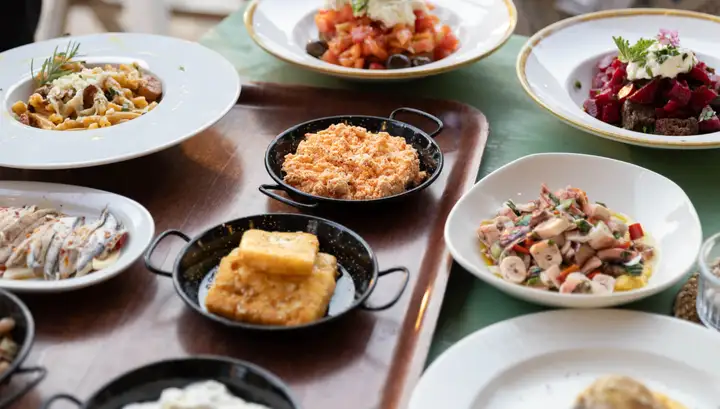 A painted table laid with plates of pasta, tomato salad, beetroot and marinated anchovies, black enamel pans of fried cheese and of a coral-coloured dip, and a bowl of chopped octopus