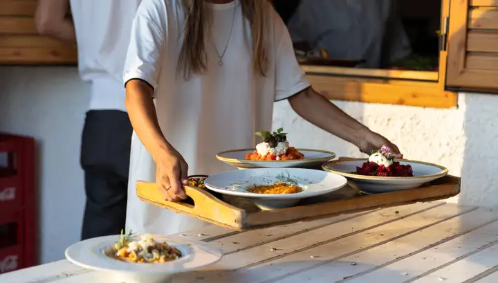 Plates of tomato salad, beetroot and a saffron-coloured dish carried out on a wooden tray to a white timber table in low sun