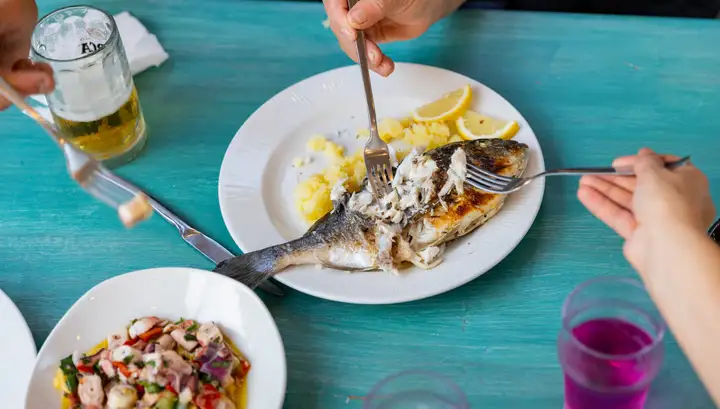 A whole grilled fish being lifted from the bone on a white plate with lemon and potato, on a turquoise painted table