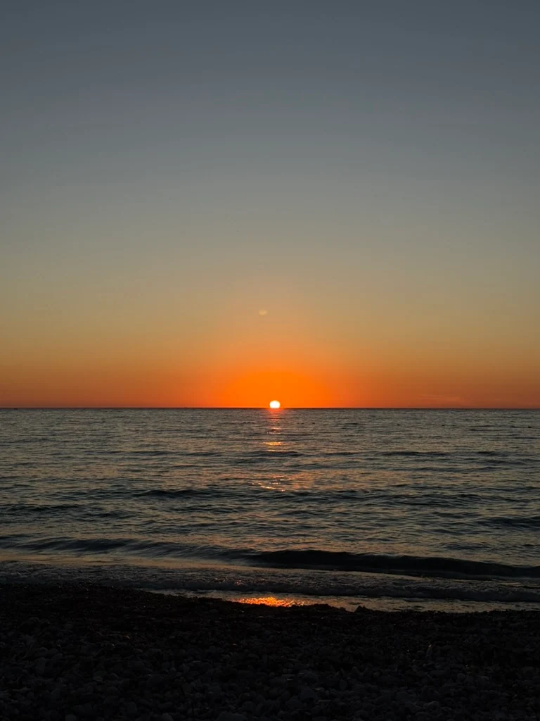 The sun touching a flat sea horizon off Spile beach, an orange band across the sky and a wet shingle foreshore in shadow