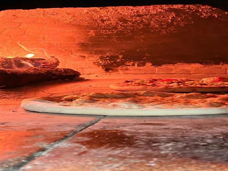 Two pizzas baking on the bare stone floor of the wood oven, embers glowing to one side