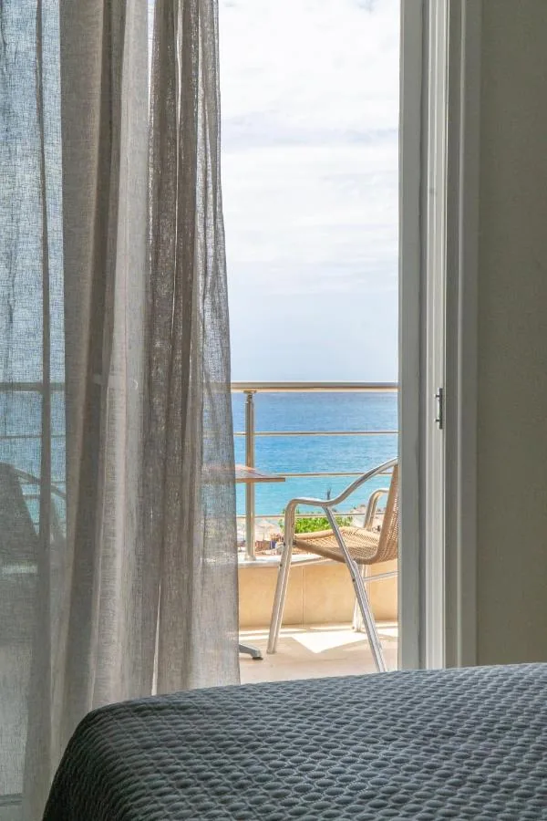 An open balcony door seen from the foot of a bed, a linen curtain drawn back, a wicker chair and the sea beyond the rail