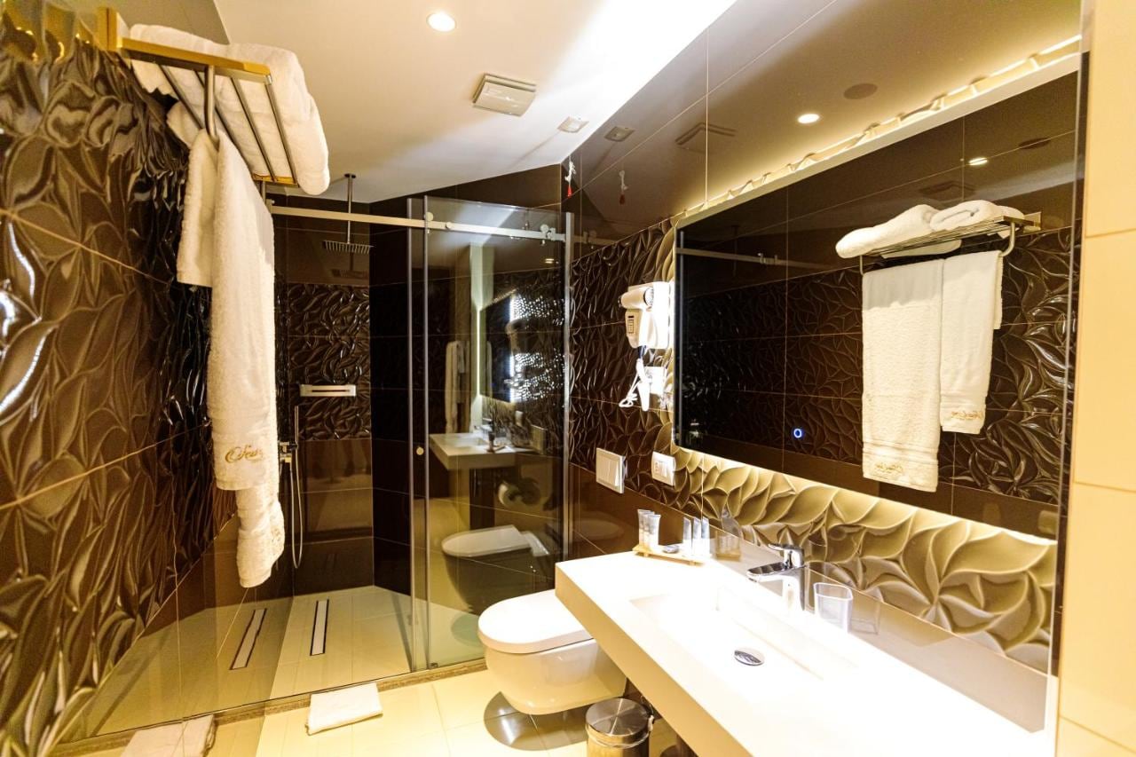 A bathroom finished in dark sculpted tile with a walk-in glass shower, a long backlit mirror, a wall-hung basin and monogrammed white towels on heated rails