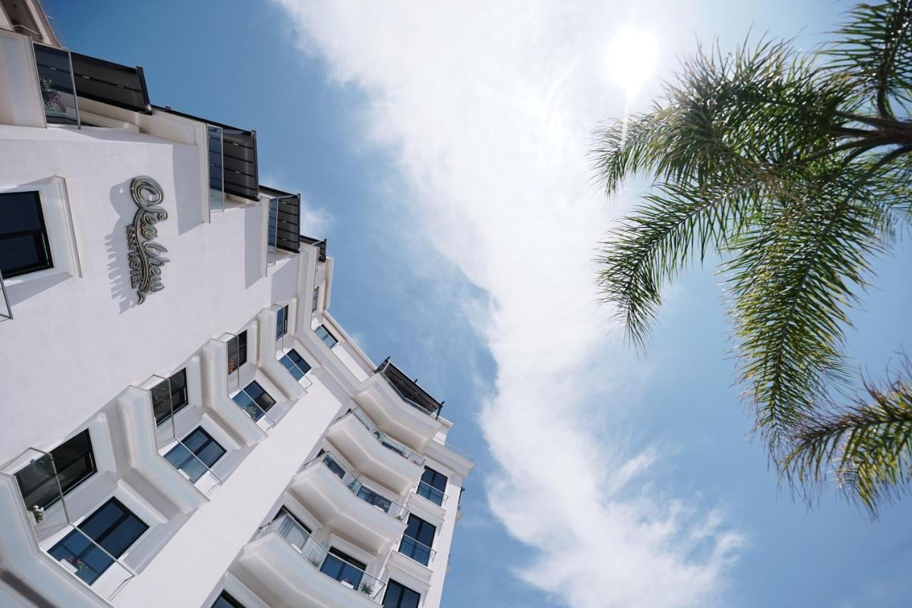 The white facade of Sea View Hotel seen from below, its curved balcony slabs stacked against a bright sky, with the hotel's script wordmark on the upper wall and a palm frond at the right