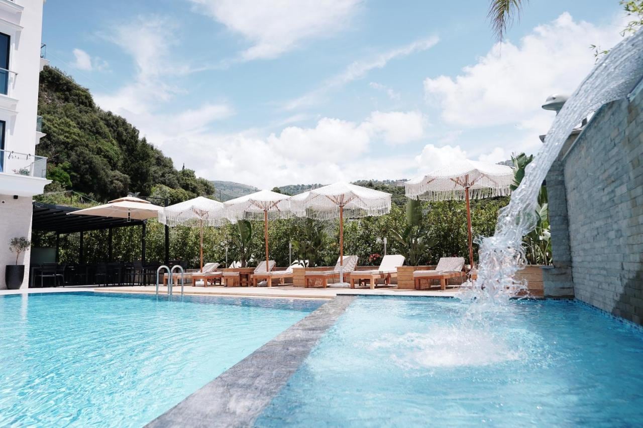 The pool at water level with a sheet of water falling from a stone wall on the right, fringed parasols and timber loungers along the far edge, and the wooded hillside behind