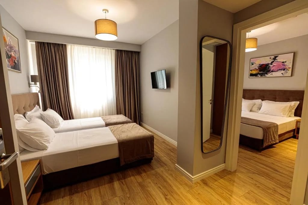 Two connected hotel rooms seen through an internal opening, twin beds with a padded headboard in the near room and a double bed in the far room, with a full-length mirror between them
