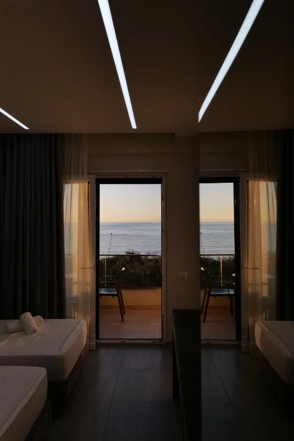 Two beds in a dim room with the sea filling both balcony doors at dusk