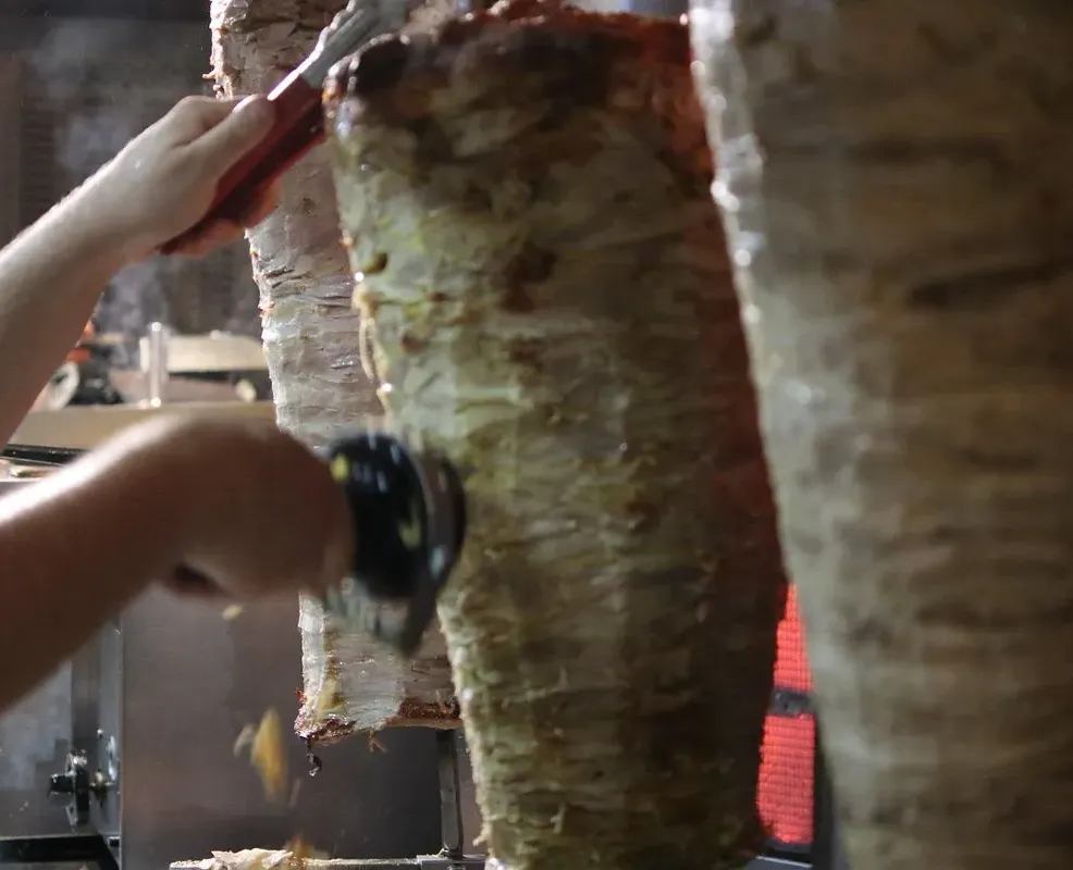 Three vertical spits of stacked meat, the left one being sliced with a handheld electric carver, a red-handled knife at the top of the middle one, a heating element glowing behind
