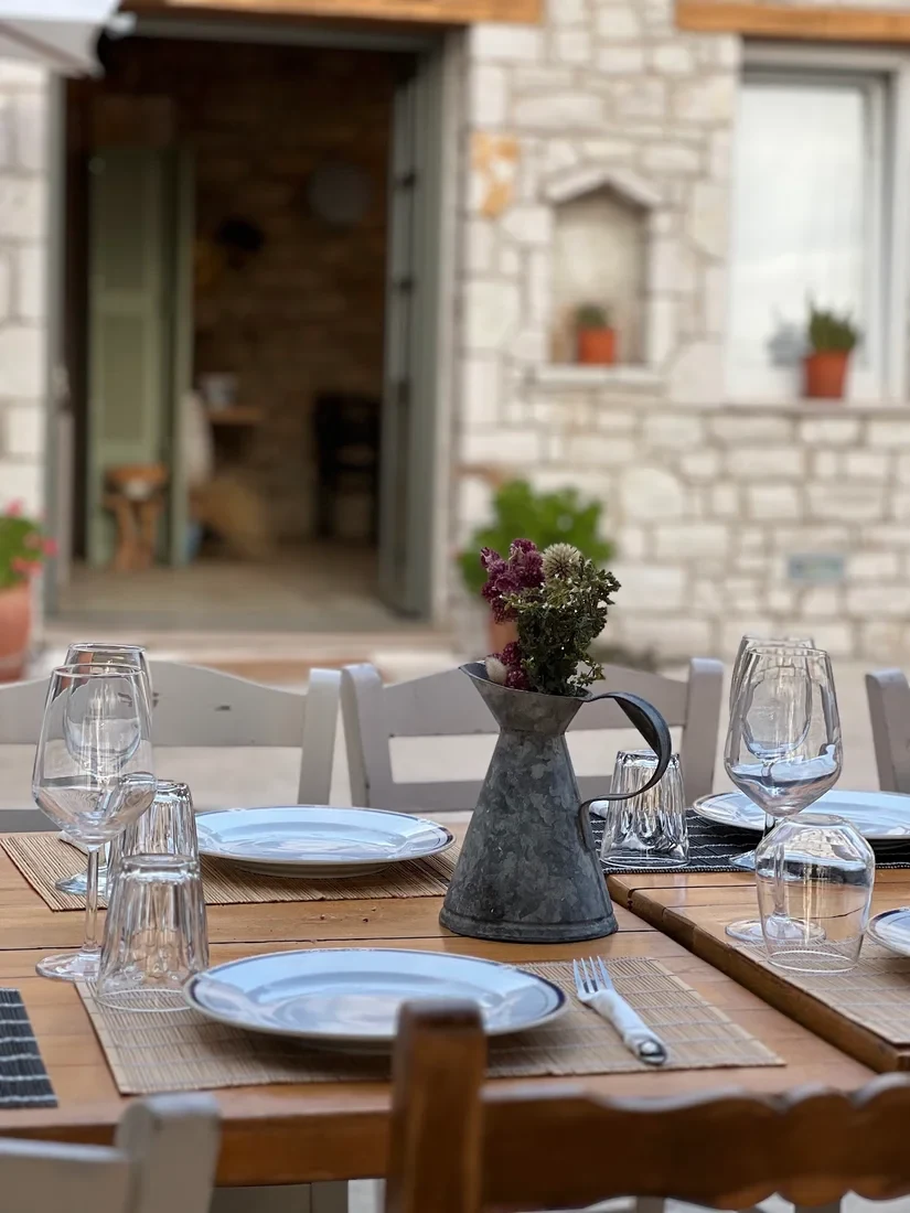 A table laid on the terrace: white plates rimmed in dark blue, wine glasses and upturned cut-glass tumblers on bamboo slat mats, a zinc jug of dried flowers in the middle, and behind it a wall of irregular pale stone with a sage-green panelled door standing open onto a stone-walled room