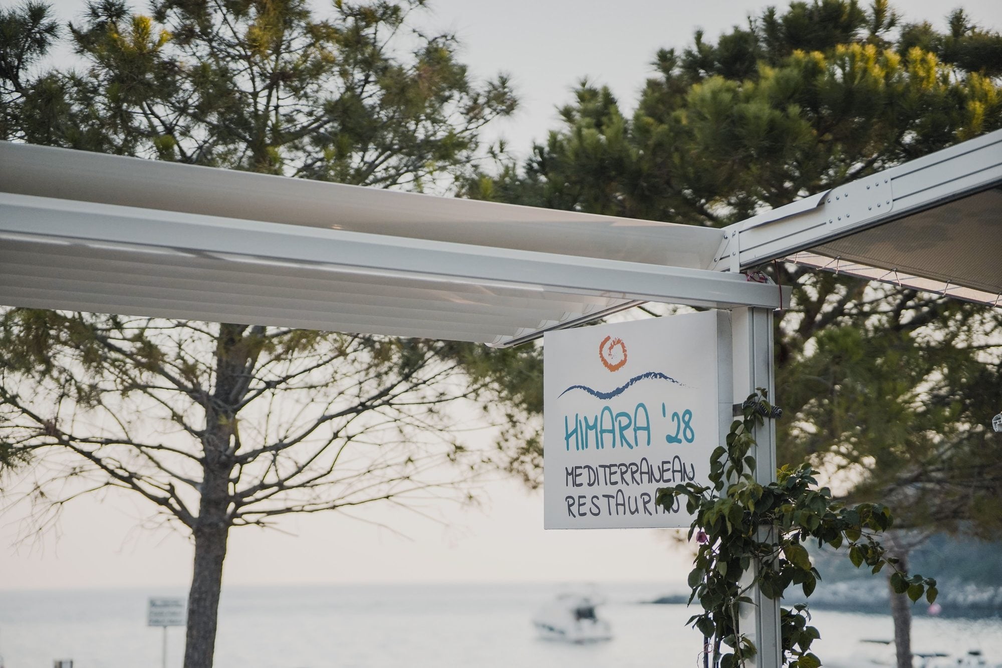 A hand-painted board hung from the awning frame, a drawn sun and wave above the lettering Himara '28 Mediterranean Restaurant, the sea and pines behind