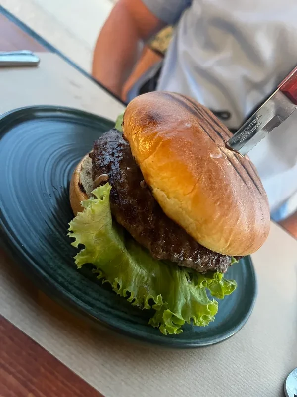 A burger in a glazed bun on a dark green plate over green leaf lettuce, a serrated steak knife pushed through the top