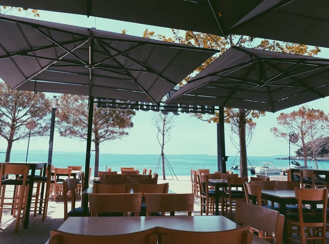 Wooden chairs and dark tables standing on beach gravel under grey canvas umbrellas, with pines, moored boats and flat turquoise water beyond