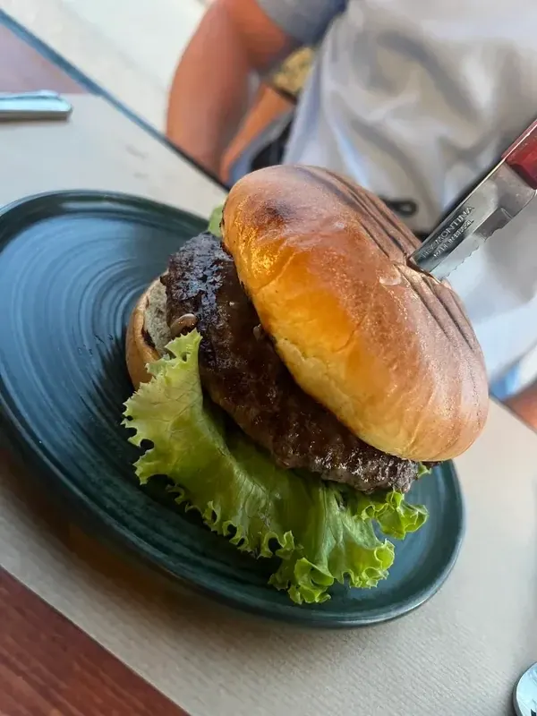 A burger in a glazed bun on a dark green plate over green leaf lettuce, a serrated steak knife pushed through the top