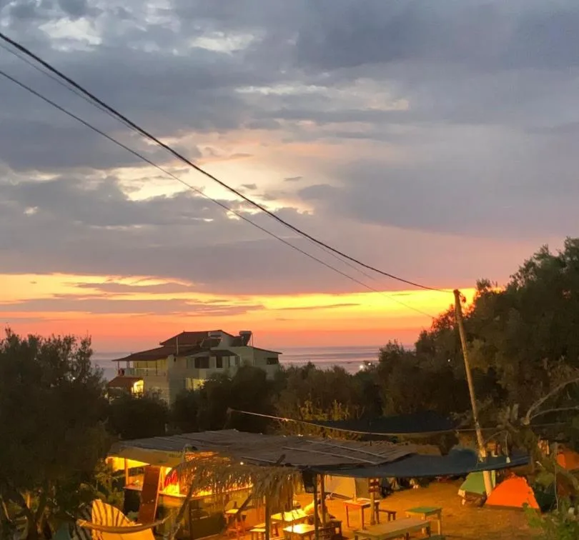 Olive Camping at dusk — the thatched servery lit warm below, tents on the pitches beyond, and the Ionian horizon under an orange sky