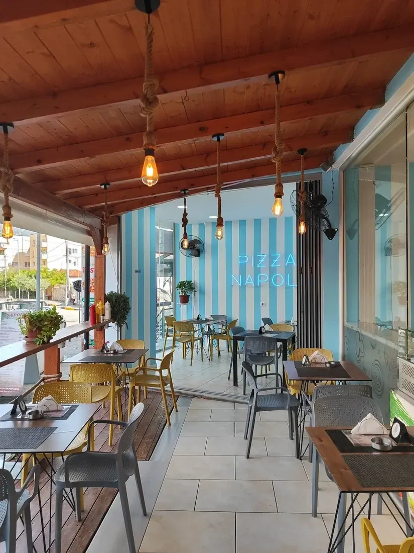 The covered terrace: a pine-boarded ceiling, bulbs hung on knotted rope, a back wall painted in wide blue and white stripes with the name across it in blue neon, and yellow and grey chairs at dark tables