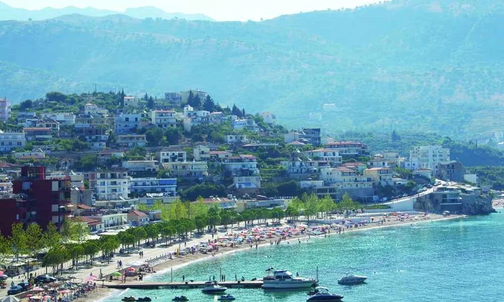 Himarë from above — the town beach, the pier and the promenade below the hillside, and the mountains behind. The town the pizzeria stands in, not the pizzeria