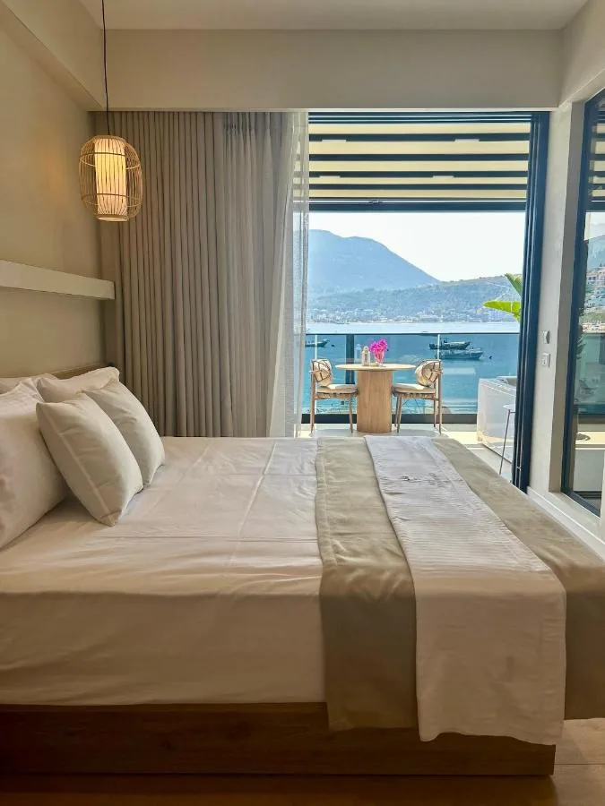 A made bed facing an open glass wall, with a balcony table for two, moored boats and a headland beyond
