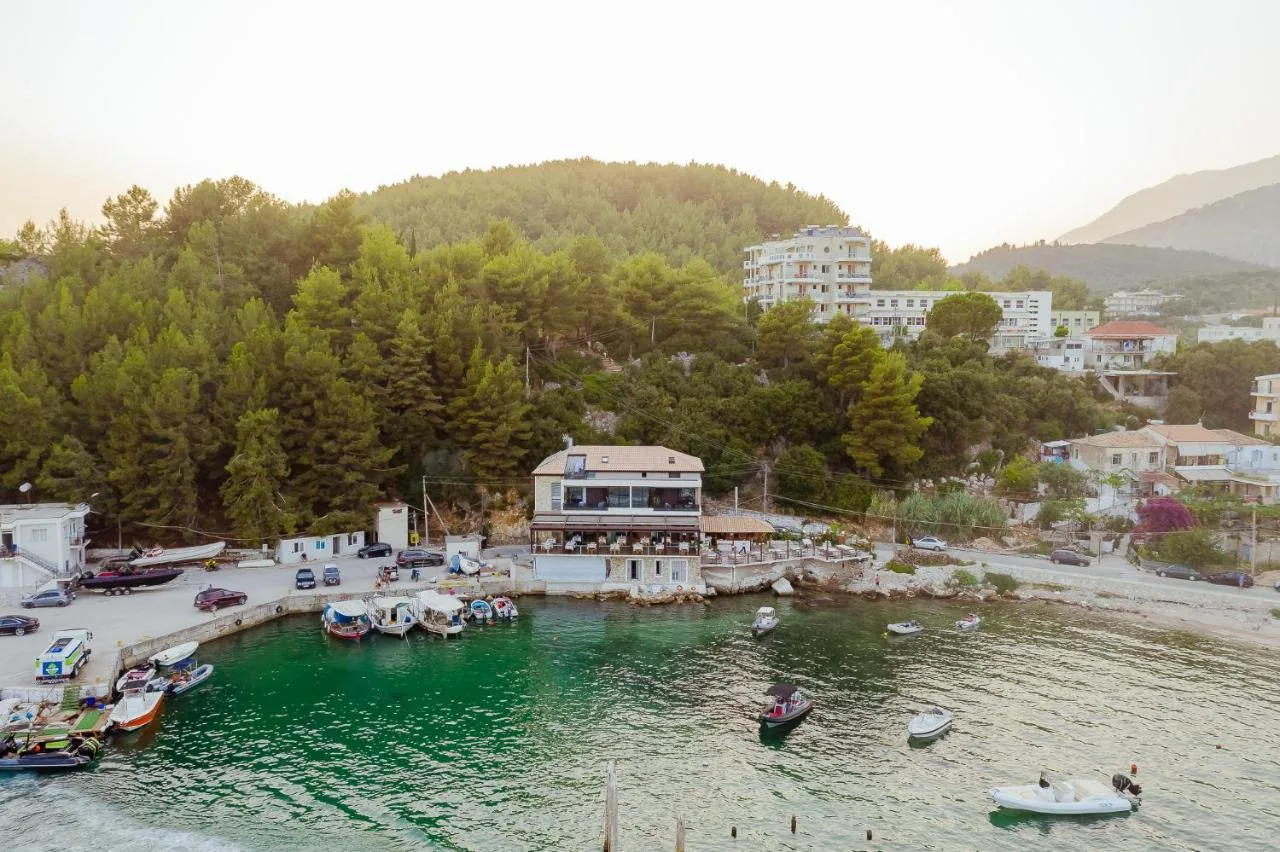 The harbour at Himarë photographed from the air at dusk, boats moored inside the mole below a wooded headland