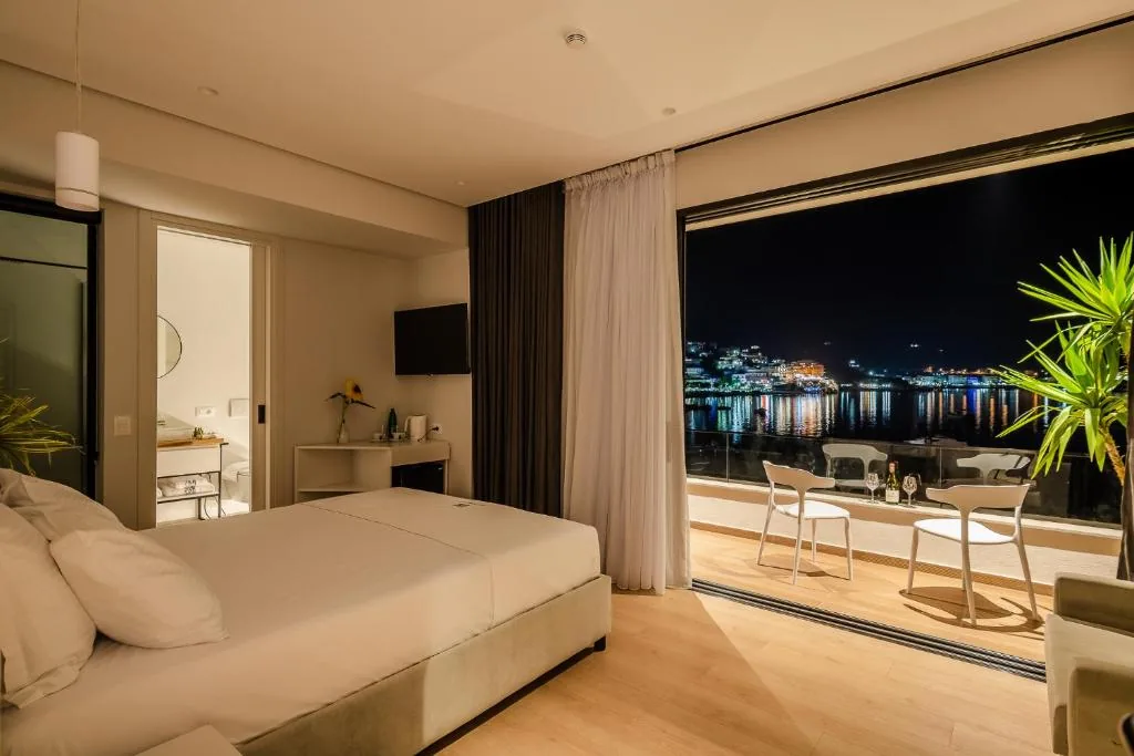 A room after dark, the glass wall open onto a lit balcony, the lights of the far shore reflected in the water