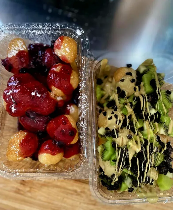 Two takeaway trays of small fried pastries, one under berry sauce and icing sugar, one under kiwi and crushed biscuit