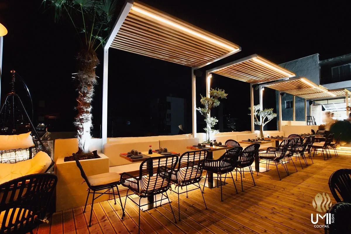 The deck after dark, three timber pergolas lit along their undersides, black rattan chairs at set tables and olive trees in the planters