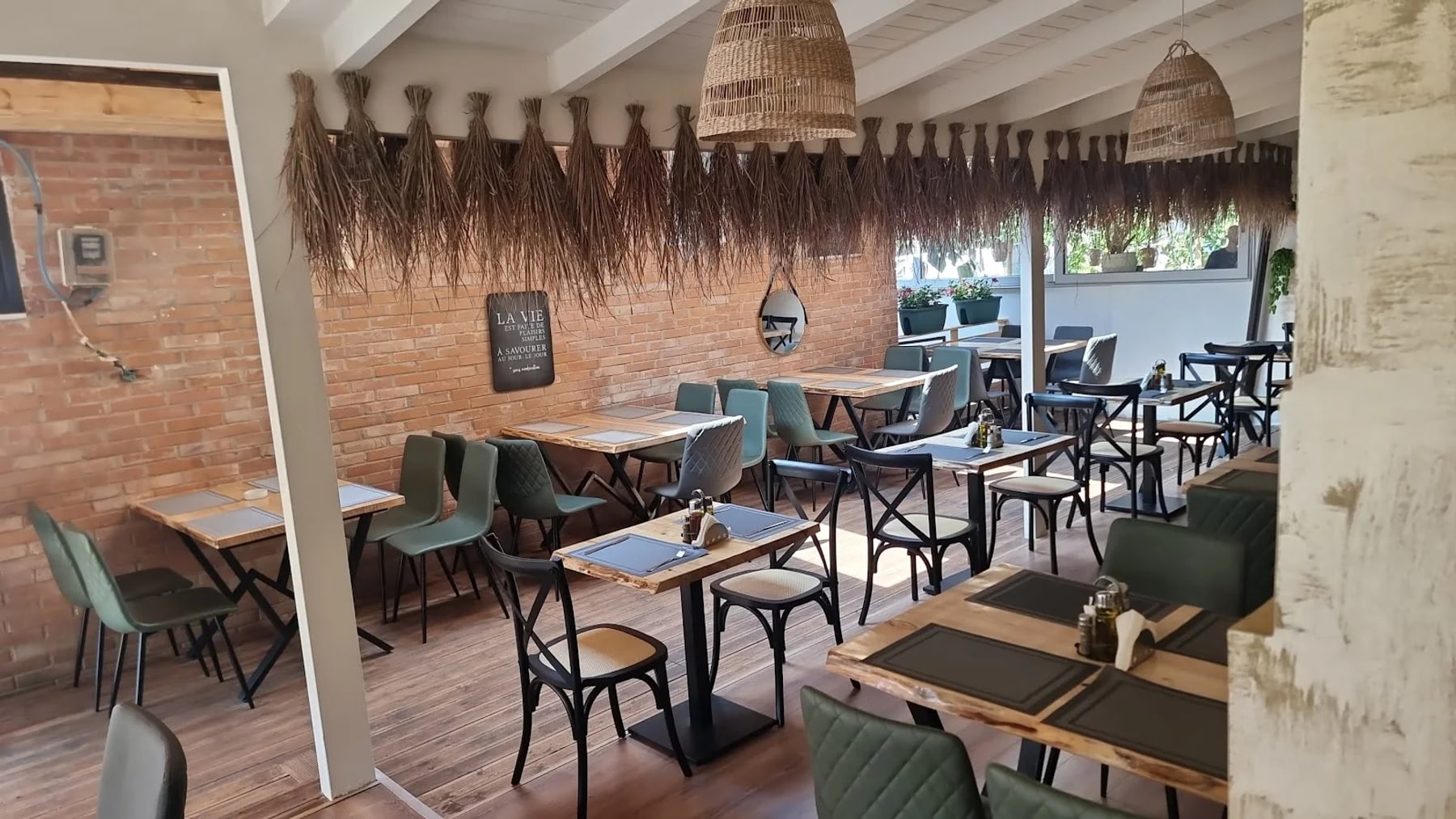 Rustic brick-walled interior with green chairs and set tables, under whitewashed rafters hung with rattan pendant shades and a fringe of dried thatch
