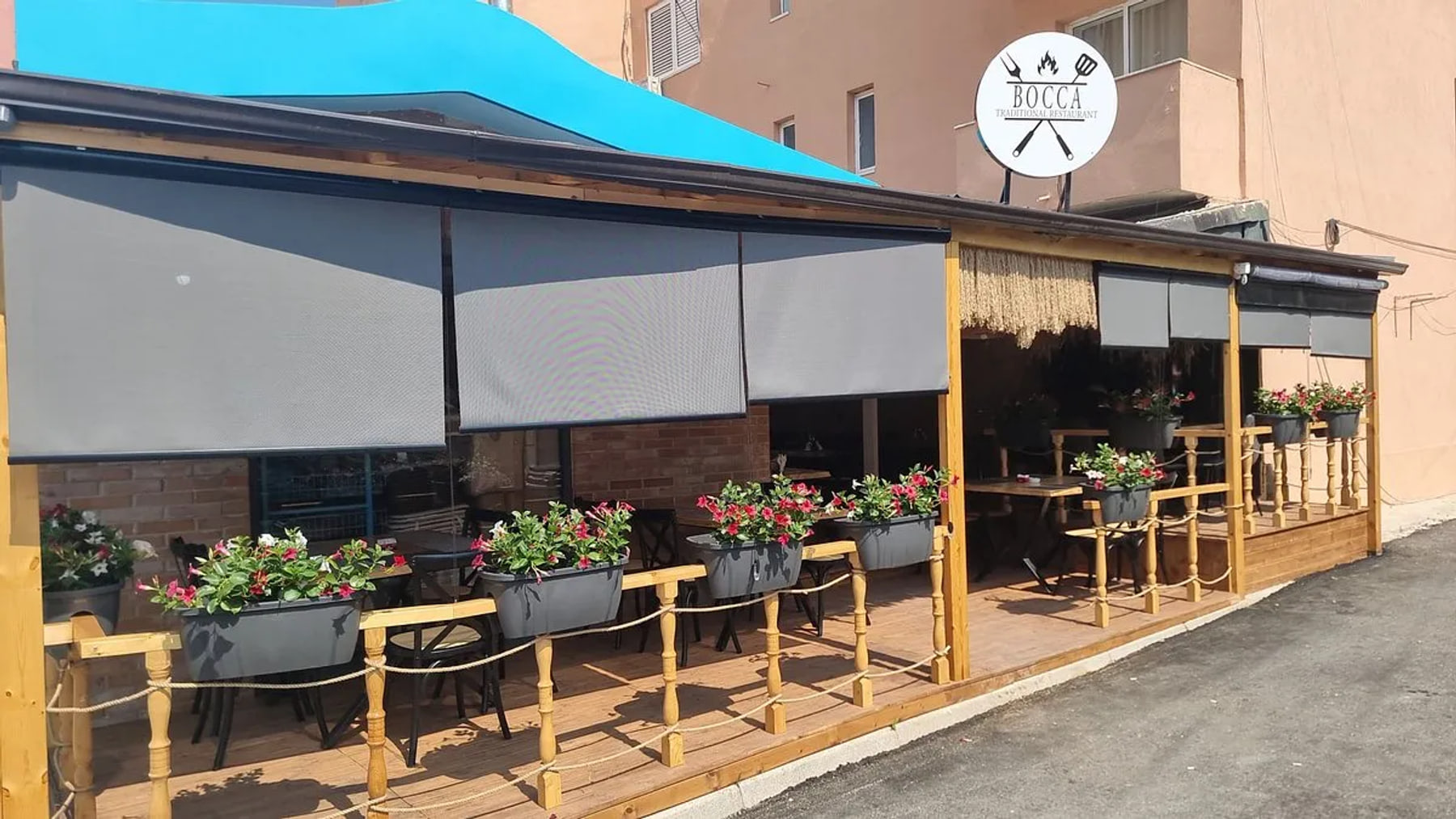 The covered timber veranda from the road, turned-wood posts strung with rope, grey planters of red and white flowers, pale grey blinds lowered over the front