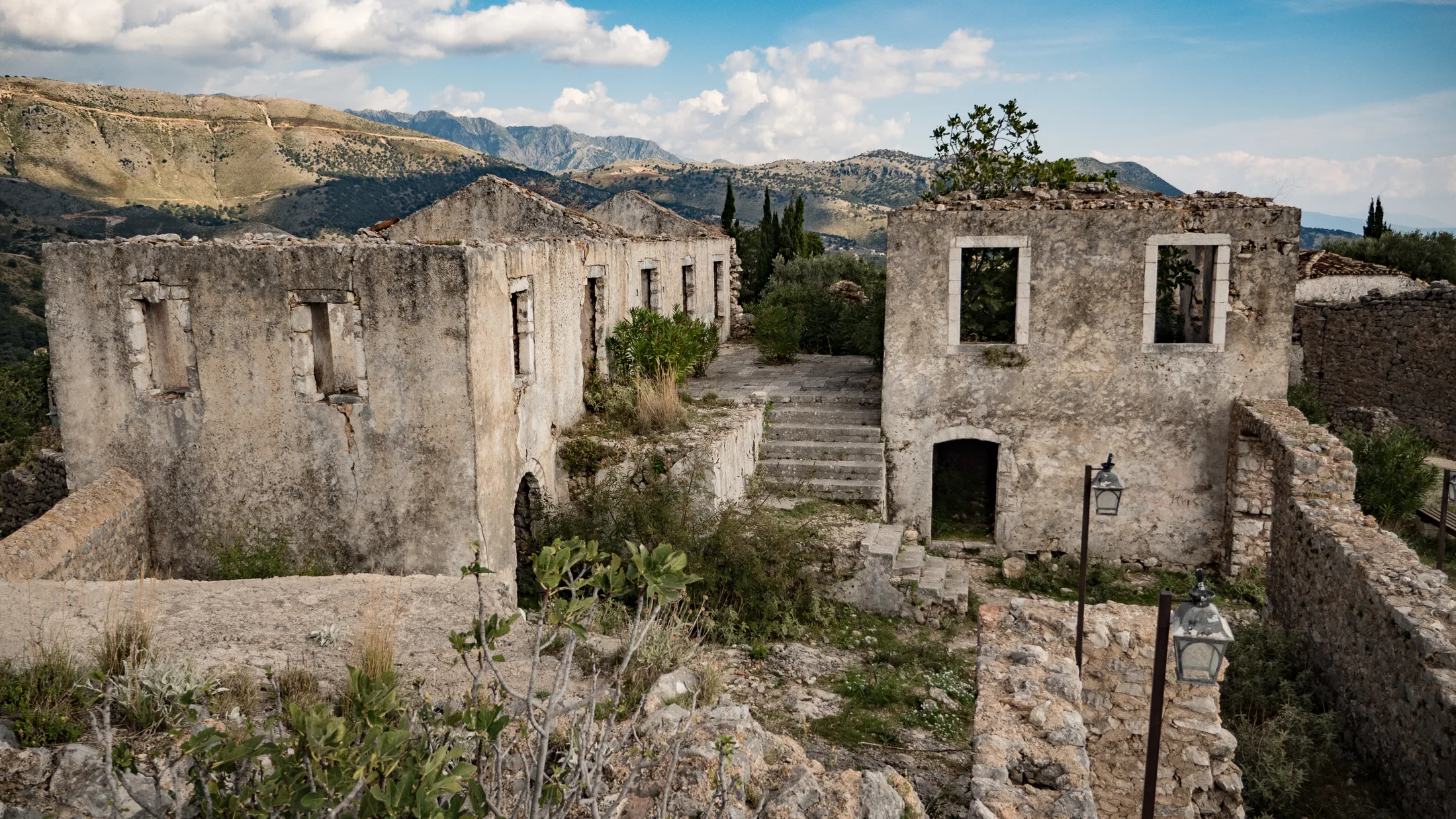Roofless stone houses in rough limestone along a stepped lane in Himarë old town, a fig growing out of one wall, dry scrub-covered limestone ridges behind