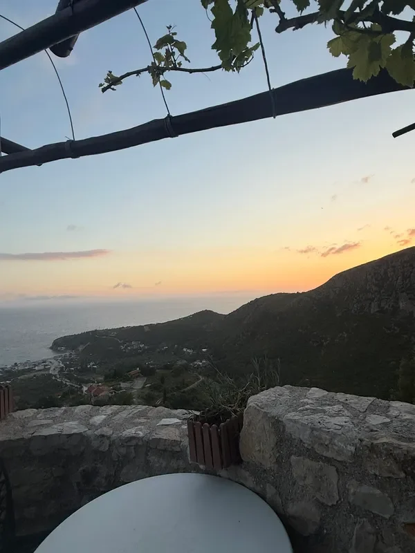 Dusk from the parapet at Café Butterfly — the sky apricot above a flat grey sea, a wooded ridge running out to the right, houses scattered on the slope below, two peeled roof poles and a vine crossing the top of the frame