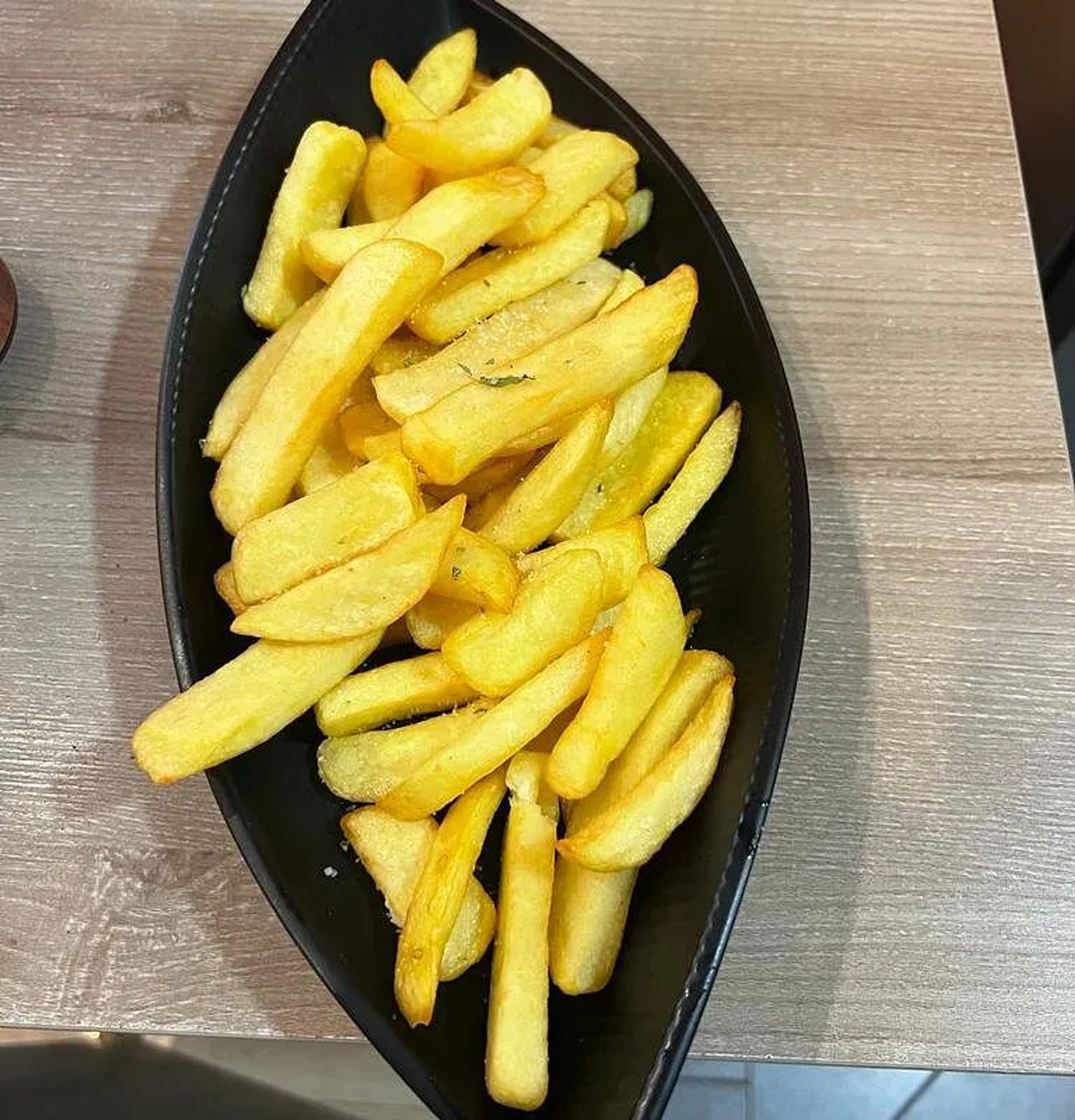 A black leaf-shaped dish of thick-cut fries, salted and flecked with herbs, on a pale timber table