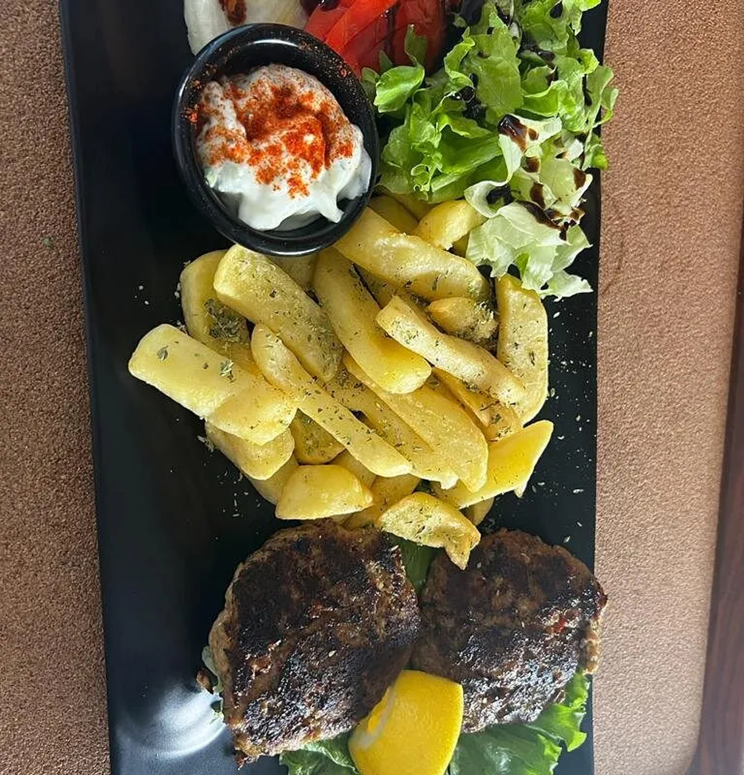 A long black plate carrying two patties taken dark, thick-cut fries dusted with herbs, dressed leaves, a tomato wedge, a raw onion segment and a ramekin of white sauce dusted red