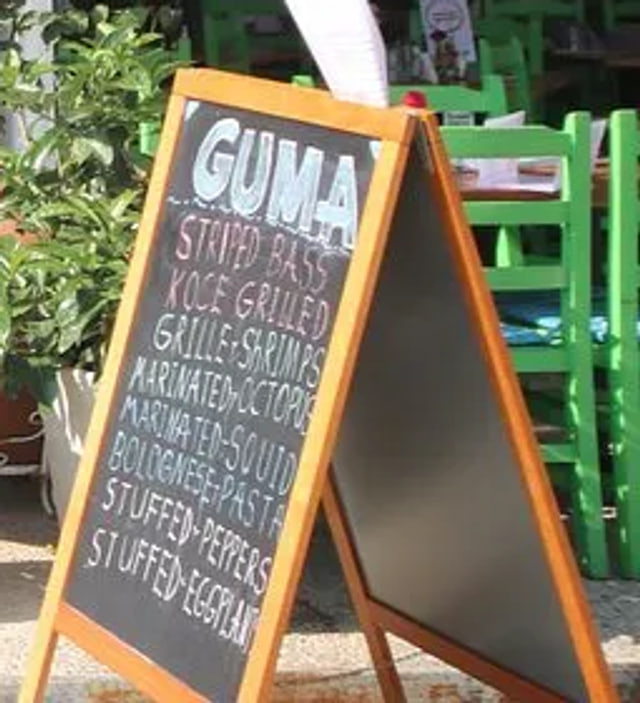 A wooden A-frame chalkboard on the pavement, GUMA chalked across the top in pale blue above a list of dishes, the first two lines in pink and the rest in white