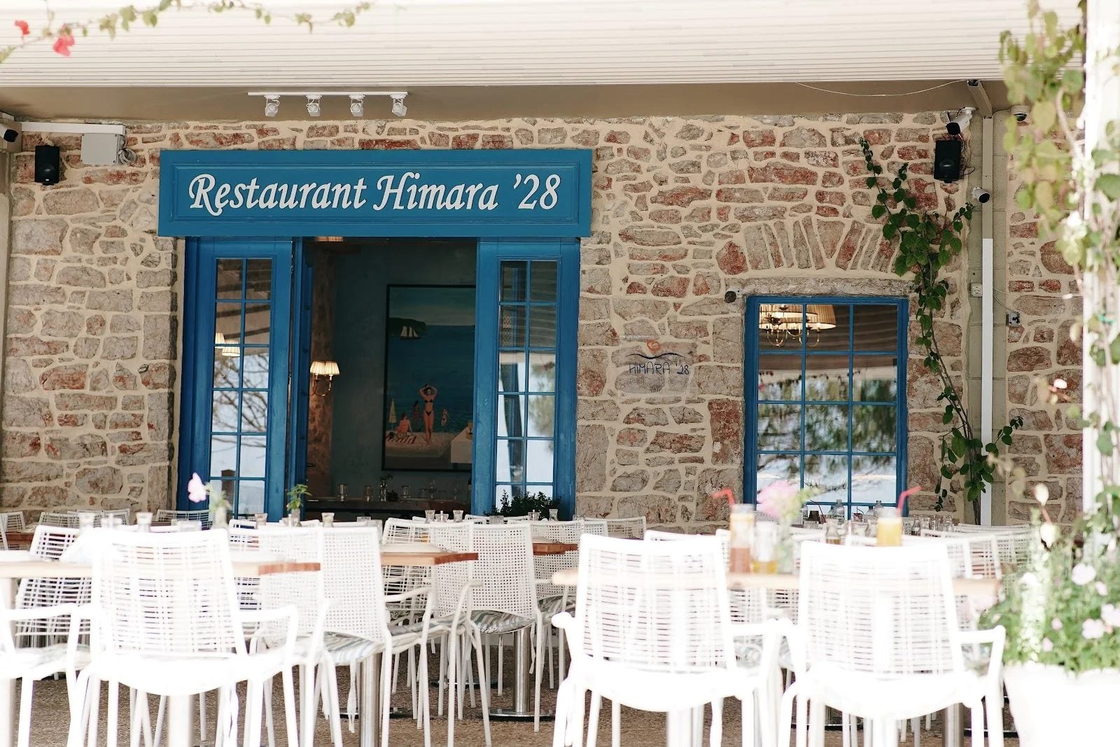 The restaurant's rubble-stone frontage with blue joinery and a blue fascia reading Restaurant Himara '28, white chairs at pale timber tables on the gravel terrace in front