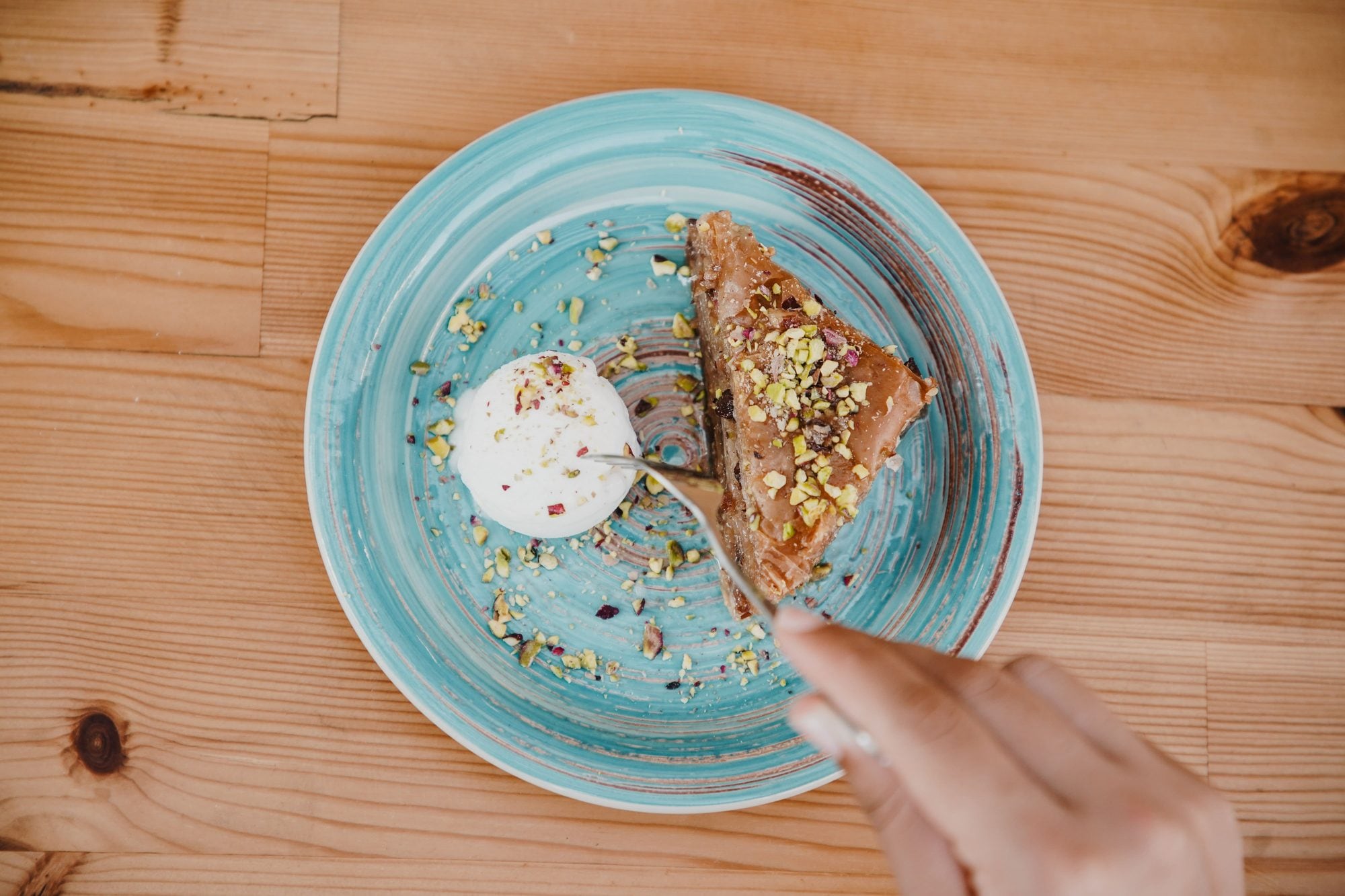 A wedge of layered pastry under crushed pistachio beside a scoop of cream, on a turquoise glazed plate, a fork coming in from the right