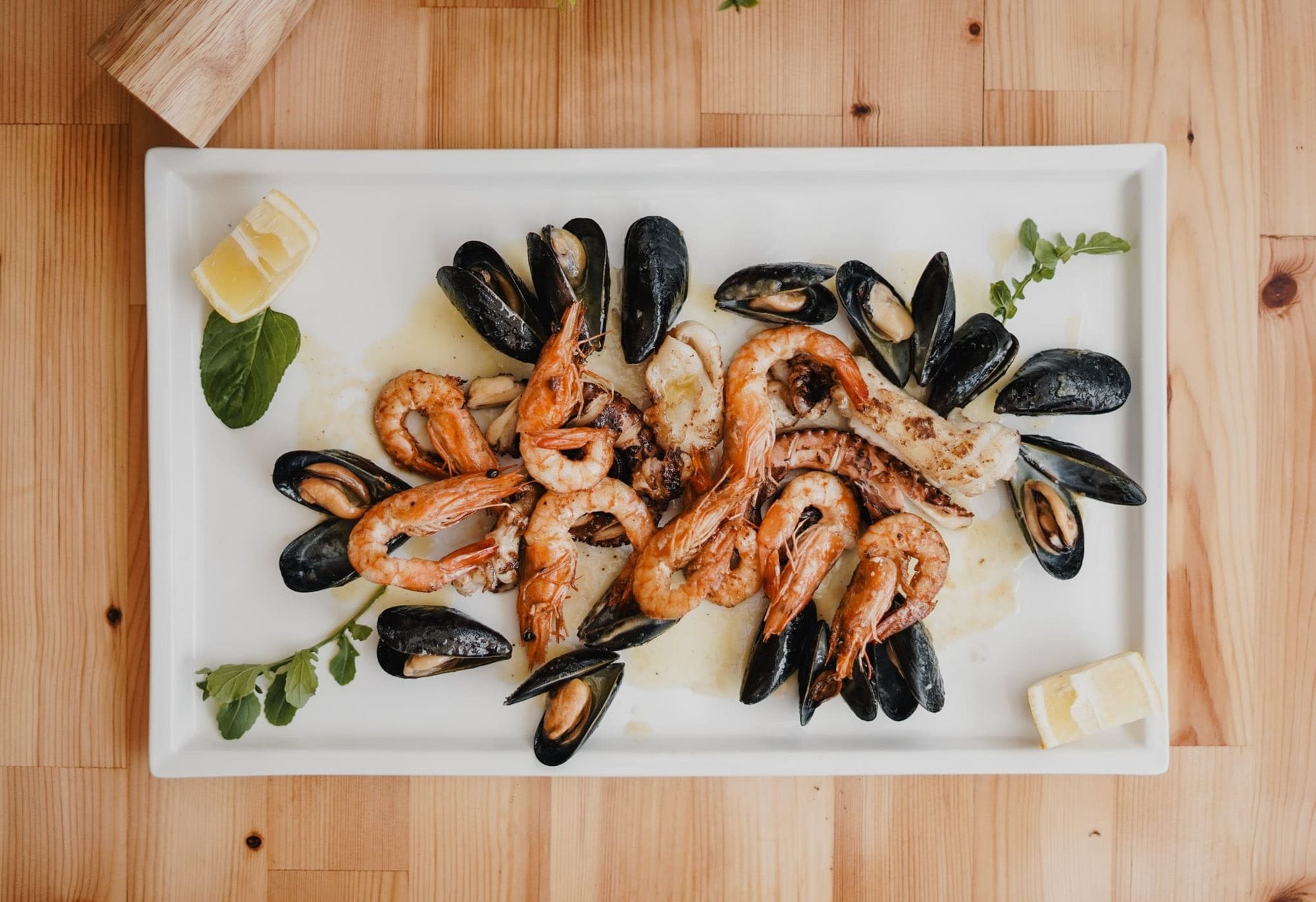 Grilled prawns, squid, octopus and black mussels arranged on a rectangular white platter with lemon wedges and green herbs