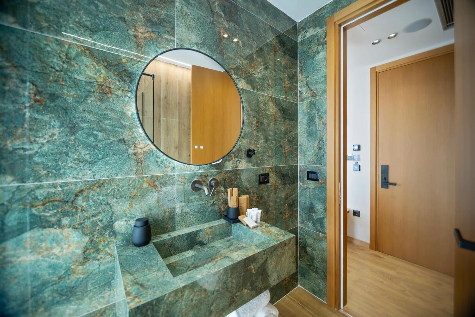 A bathroom at Hotel Potami lined in green marble, with a round black-framed mirror above a basin and counter built from mitred panels of the same green stone
