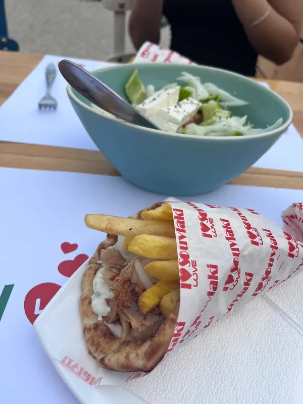 A gyros wrap in I Love Souvlaki's own printed paper, chips inside the pita, a bowl of salad with feta behind it