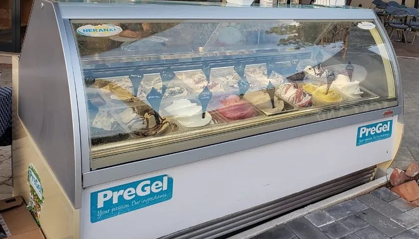 A curved-glass gelato cabinet standing on the pavement outside, its pans scooped, pale blue tags standing in them