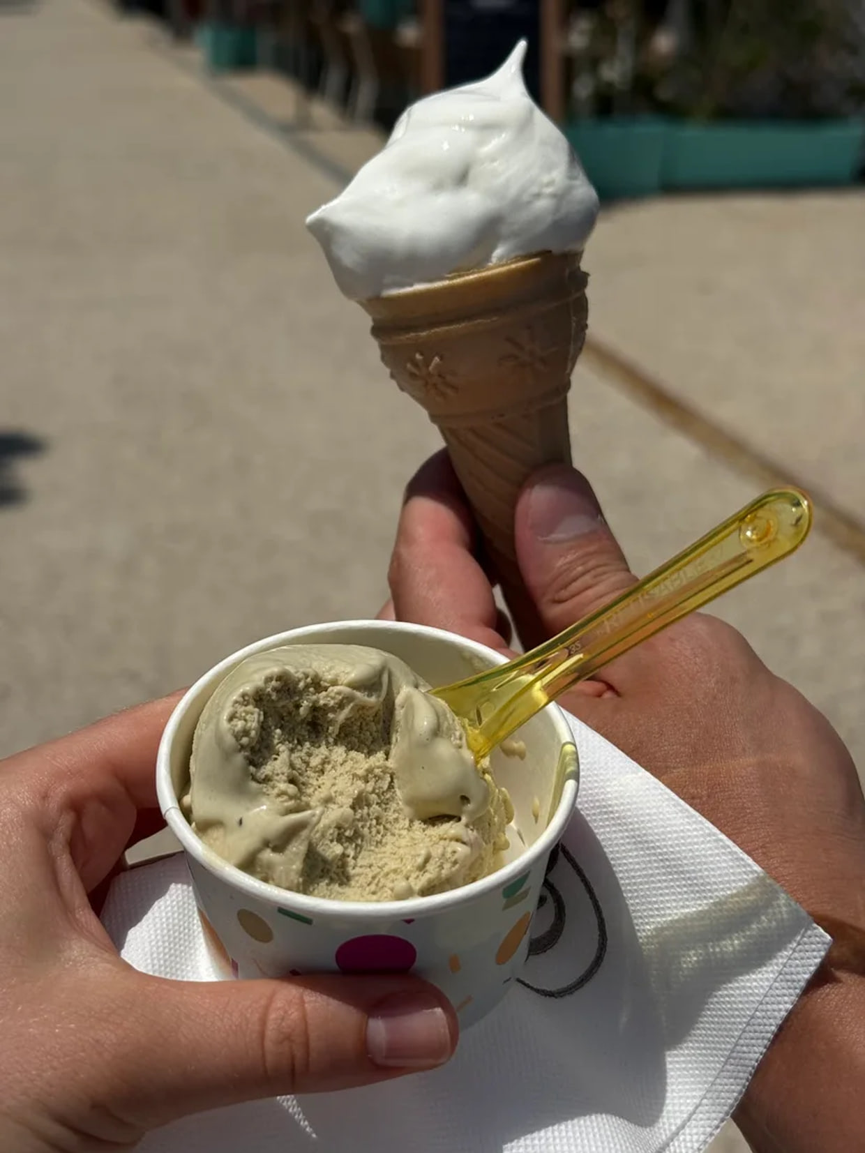 A soft-serve swirl on a wafer cone held in one hand and a cup of scooped gelato in the other, out on the street