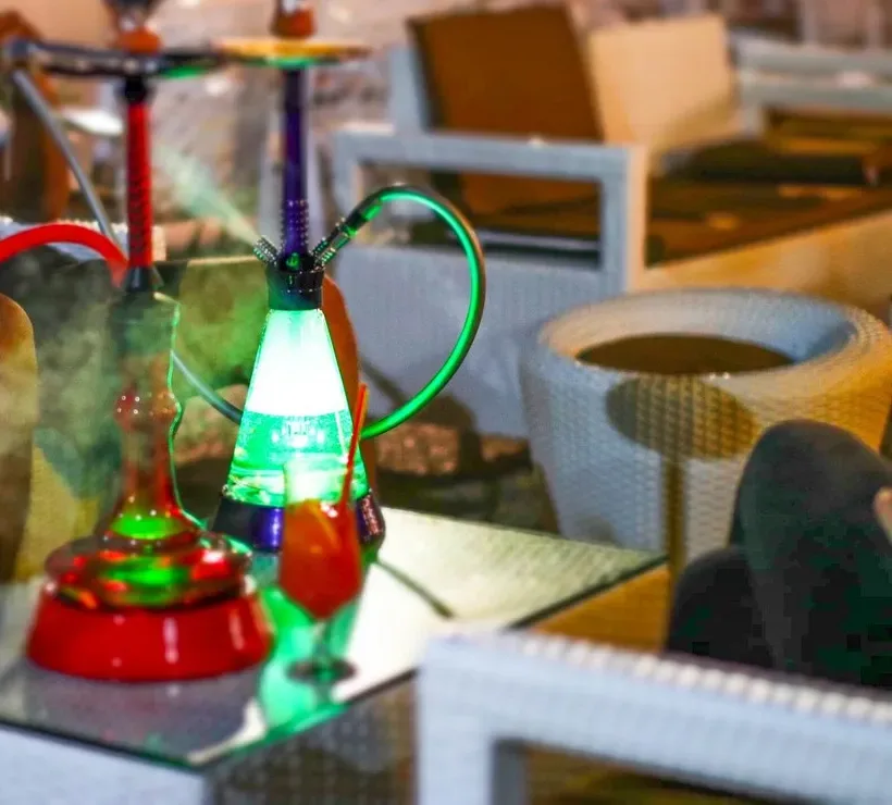 Two shisha pipes with illuminated bases on a low glass-topped table on the terrace at Lido Lounge, a cushioned sofa and a woven side table behind