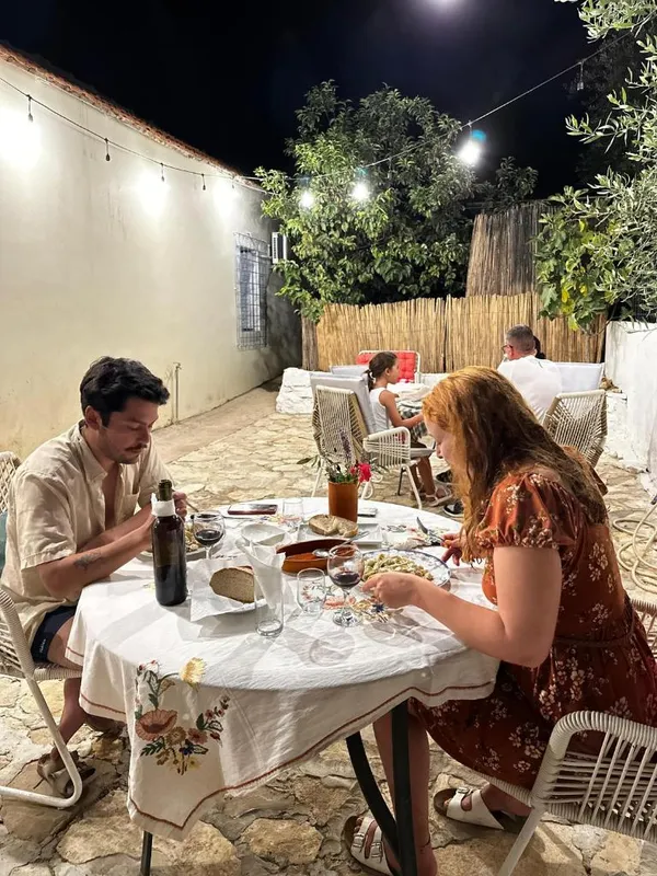 Guests eating at a table in a walled courtyard at night under a string of lights, with a bamboo screen and an olive tree behind them