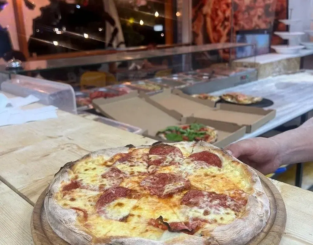 A round pizza on a wooden board at the counter: cheese under salami, its raised rim flour-dusted and spotted dark where it caught the heat, with trays and open flat boxes on the counter behind