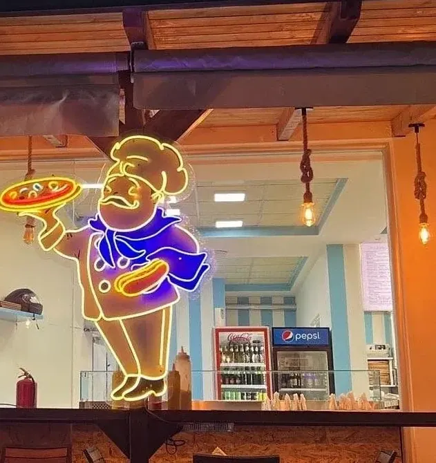 The cook in neon over the counter — a figure outlined in gold, in a buttoned chef's jacket and hat, blue neckerchief, carrying a pizza flat on one hand — with the rope-hung bulbs and the blue and white striped wall behind