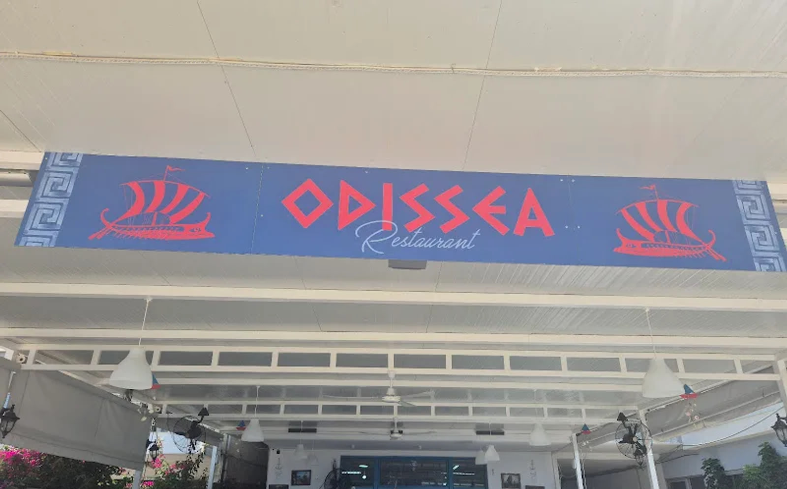The painted Odissea Restaurant sign, red lettering and two galleys on a blue board with a Greek key border, fixed under the white pergola roof