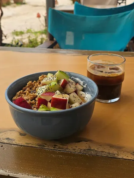 A granola bowl with apple, kiwi and dragon fruit beside an iced coffee on a painted wooden table