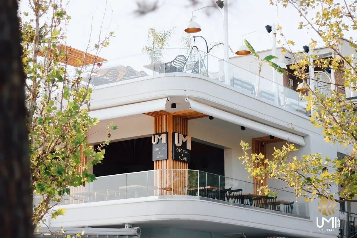 The white corner building that carries UMI, its two black signs reading Cocktail & Sushi between timber columns, plane leaves across the foreground